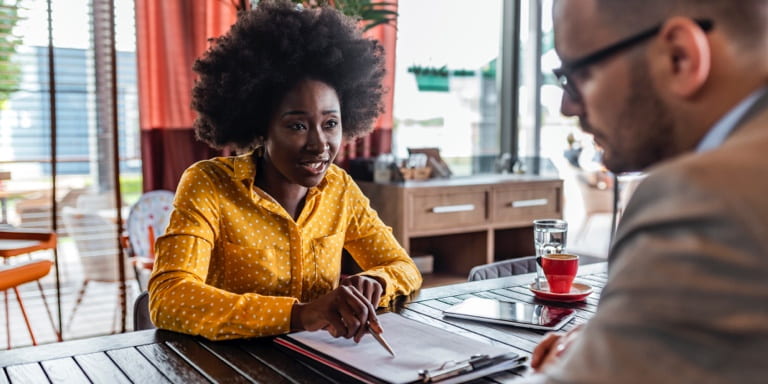 A woman and her male colleague having a business discussion at a table, about some documents in a file.