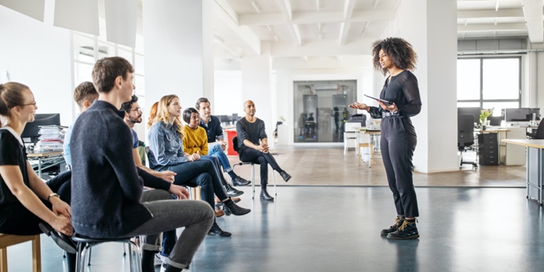 A woman in dark gray pants and a dark gray top, giving a presentation to a group of people seated before her in a modern, open plan office building.