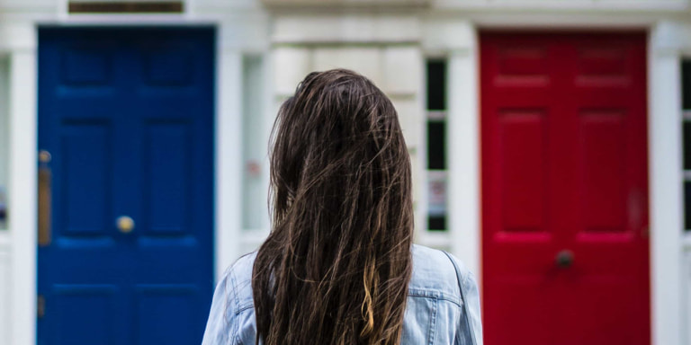 A back shot of a woman with long brunet hair facing three doors, the left one is blue, the middle is white and the left is red.