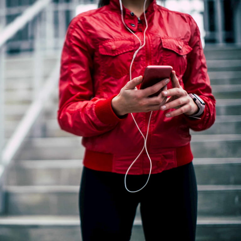 Leading a healthy lifestyle, an athletic lady in a red sporting gear, holding her cell phone with earphones in her ears with stairs in the background.