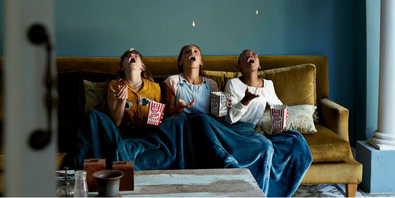 Three girls sitting and eating popcorns on a couch with a blanket in a home bought with the help of FundsAtWork's housing loan guarantee.