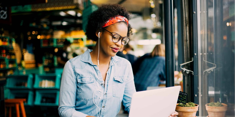 A lady wearing a denim shirt, glasses and a red hair band with earphones on, sits in a cafe with an open laptop in front of her.