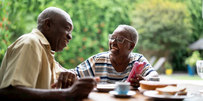 An elderly couple having afternoon tea, smiling and talking to one another lovingly. The woman embraces her husband by touching him on his arm.