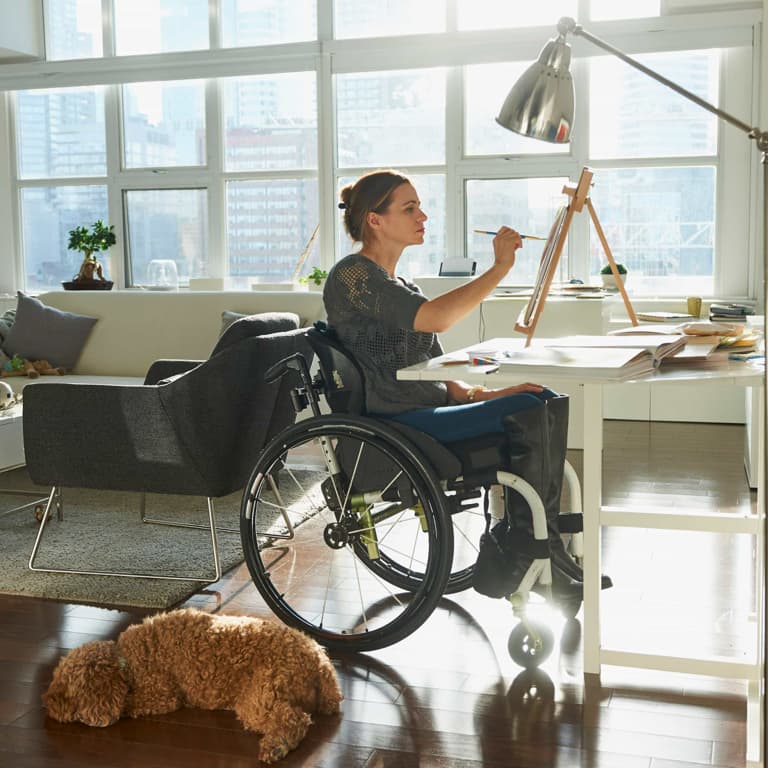 A young lady in a wheelchair sitting at her drawing desk, painting on an easel. She is in her modern apartment with lots of natural light and her dog is lying on the floor beside her.
