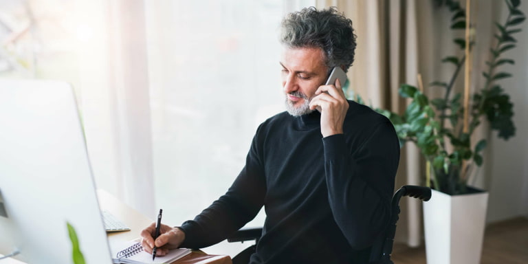 A middle-aged man sitting in a chair near a window. He is writing down a message from a phone call he is taking.