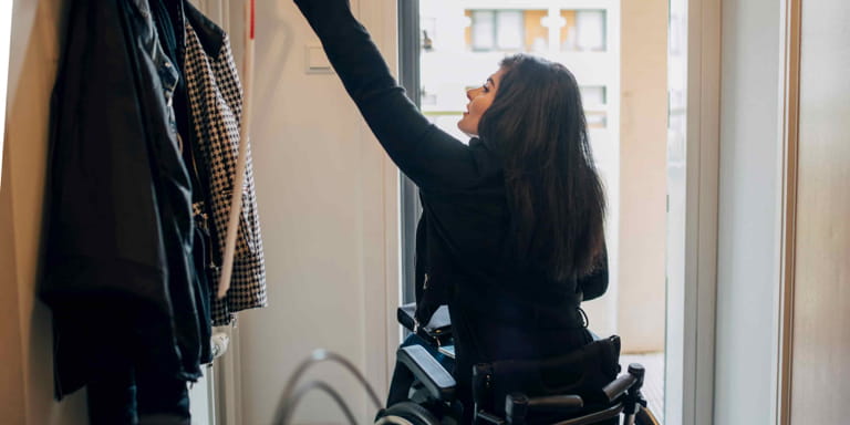 A young female wearing black is sitting in a wheelchair reaching up to grap her coat from the rack as she is about to leave the building.