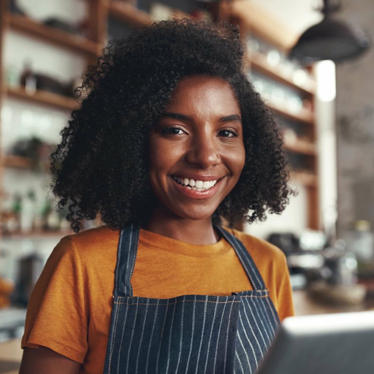 A young black female with a big smile wearing a mustard t- shirt and a striped denim apron standing in front of shelves.