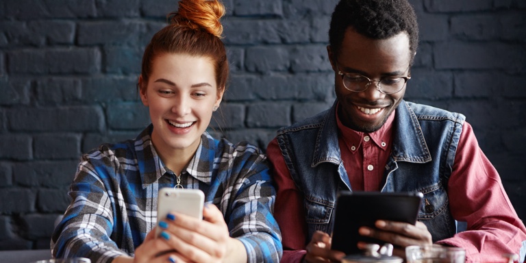 A couple of people drinking tea while looking at their cell phones smiling.