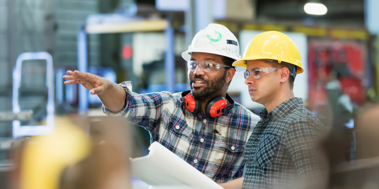 Two men wearing hard hats and safety goggles, standing in a factory discussing building plans.