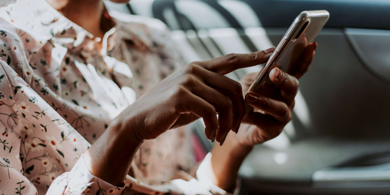 A woman in a sitting on the backseat of a car is typing on her cell phone.