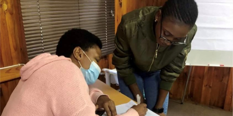 masked woman seated helping hunched man in glasses fill in a form.