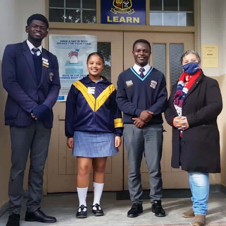 Three black children of different ages, dressed in school uniform, standing in front of the school office door with their female teacher