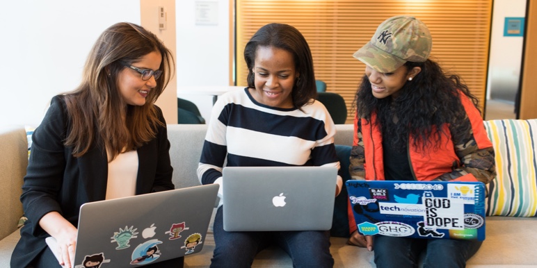 Three young woman doing on their laptops smiling. One has on a hat.