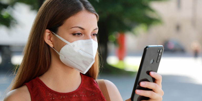 A young woman wearing a mask over her nose and mouth looks at her cell phone to check what her medical aid covers for COVID-19 related tests.