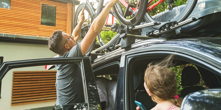 A dad carefully placing a bicycle on the roof rack while his daughter watches him from the front passenger window.