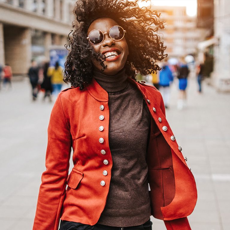 A young woman with curly hair and wearing a red jacket and sunglasses walking in a street.