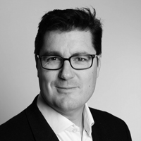 A black and white head and shoulder picture of the author of the internations diversification article, Greg Davies, in a black suit jacket and white shirt.