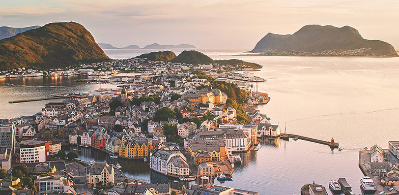 An aerial view of a coastal town in Norway, with old orange and white Norwegian style buildings with sharp, black roofs surrounded by mountains and seawater. 