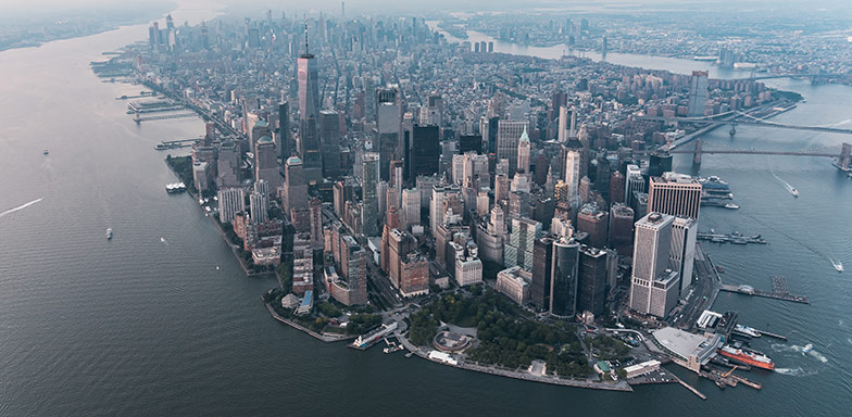 An aerial view of parts New York city and its skyscrapers surrounded by seawater and a few boats and ships around the shoreline.