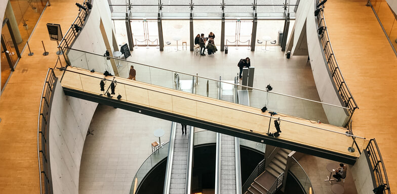 A view from the top, looking down the inside of an office building with brown laminate floor, white walls and escalators at its center.