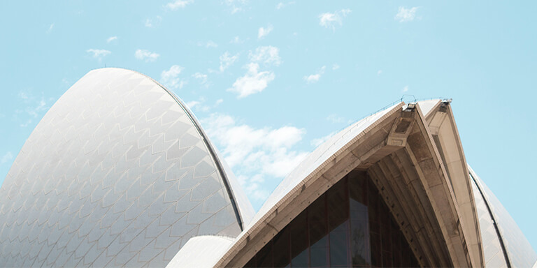 The Sydney Opera House, shot from below, to show its sharp, granite tiled roofs against a litght blue sky.
