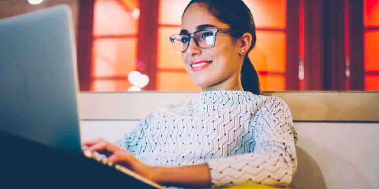 Lady wearing a white top with black dots sitting on a sofa typing on the open laptop.