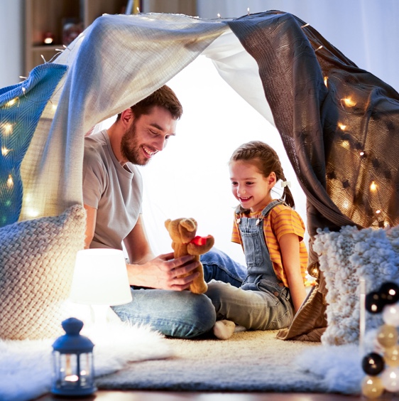 Father and his young daughter sitting under a makeshift tent in their living room. They're smiling while the father holds a small teddy bear while telling her a story.