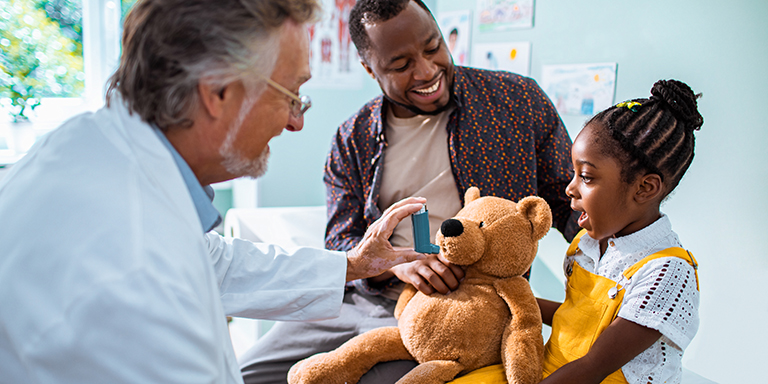 Little girl holding her teddy bear while the doctor pretends to give a metered dosage from the asthma pump so the little girl doesn’t feel scared to use the pump when she needs it.