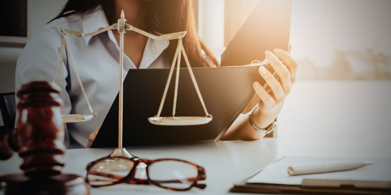 A young female lawyer reading a legal document with a pair of reading glasses, gavel and justice scales on the table in front of her.