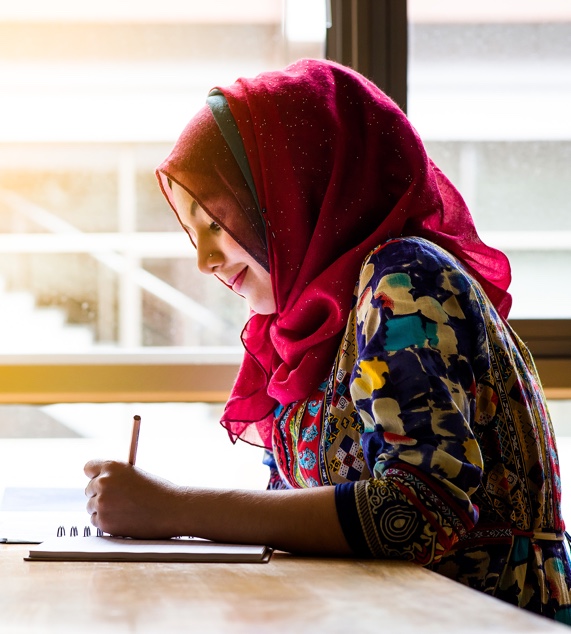 Young lady wearing a headscarf and a colorful dress sitting at a table and writing in a book. She is smiling knowing her estate administration will be handled professionally.