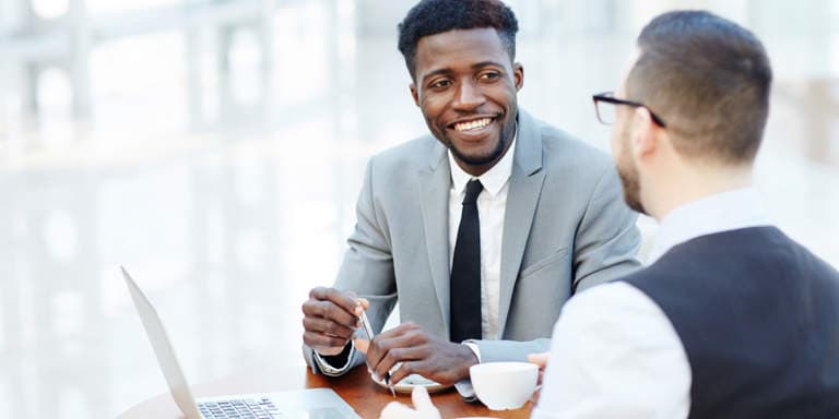 Two men sitting at a table talking and having coffee with an open laptop in front of them.