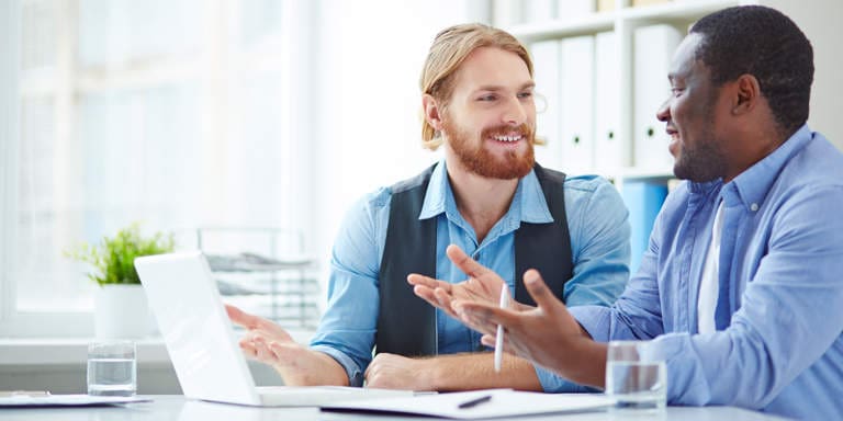 Two guys sitting at a table having a conversation with a lap top in front of them. 