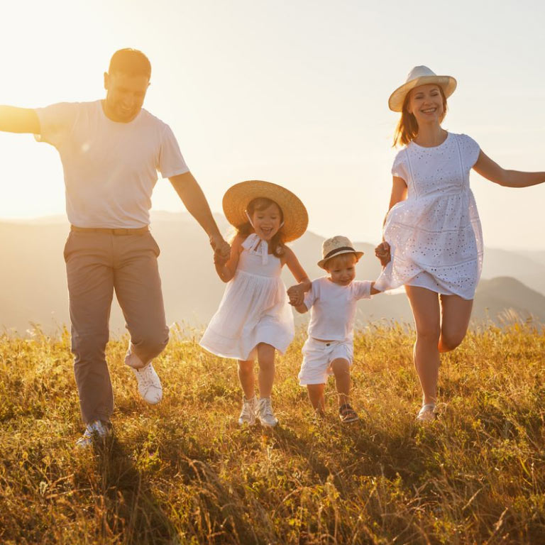 A family of four, mother and father holding their two children’s hands as they run in an open field as the sun sets.