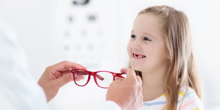 A cute young girl smiles while an optometrist holds out red-framed glasses for her to try on.