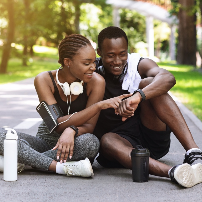 Young fit couple in gym gear sitting on the street, smiling, and looking at the young man’s fitness watch that’s tracking health activity for Momentum Multiply points.