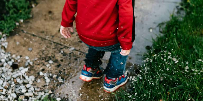 A toddler in a red jacket and blue and red sneakers jumping into a small puddle on the pavement and making a splash.