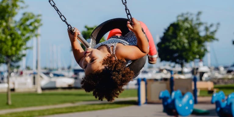 Little girl in the sunshine on a tyre swing in the park, with boats on the sea in the background.