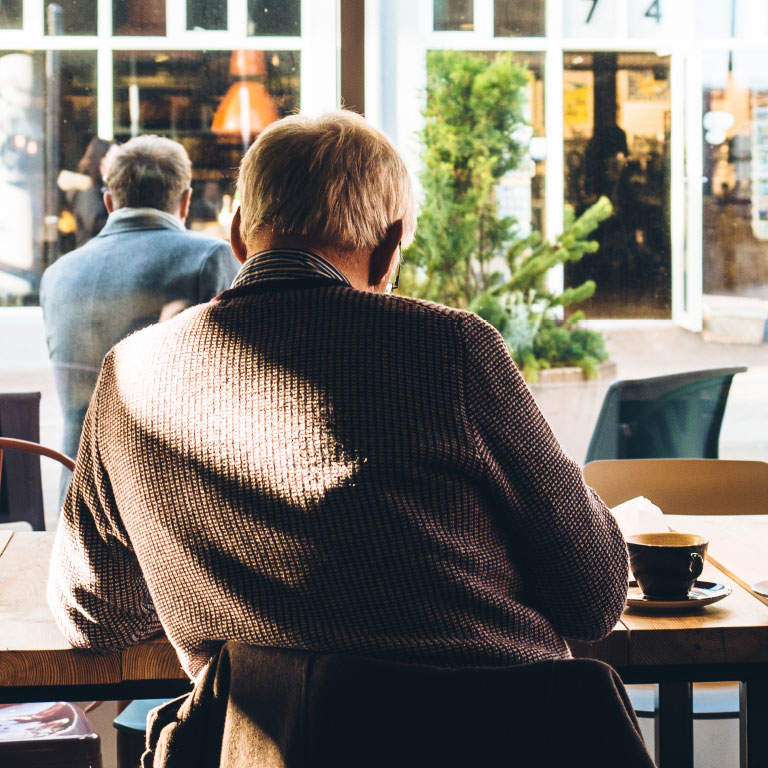 Elderly man sitting at a table in café drinking a cup of coffee and looking out the window with a view of the shops in front of him.