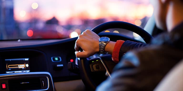 A businessman drives a luxury car in busy traffic at dusk. He is resting his hand on the steering while and you can see his expensive watch.