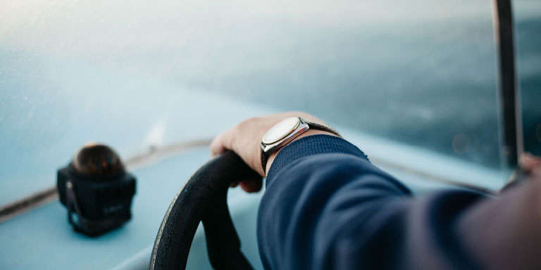 Close up of a man’s arm resting on the steering wheel of his car with a camera on the dashboard. He is wearing a watch and long-sleeved blue suit jacket.