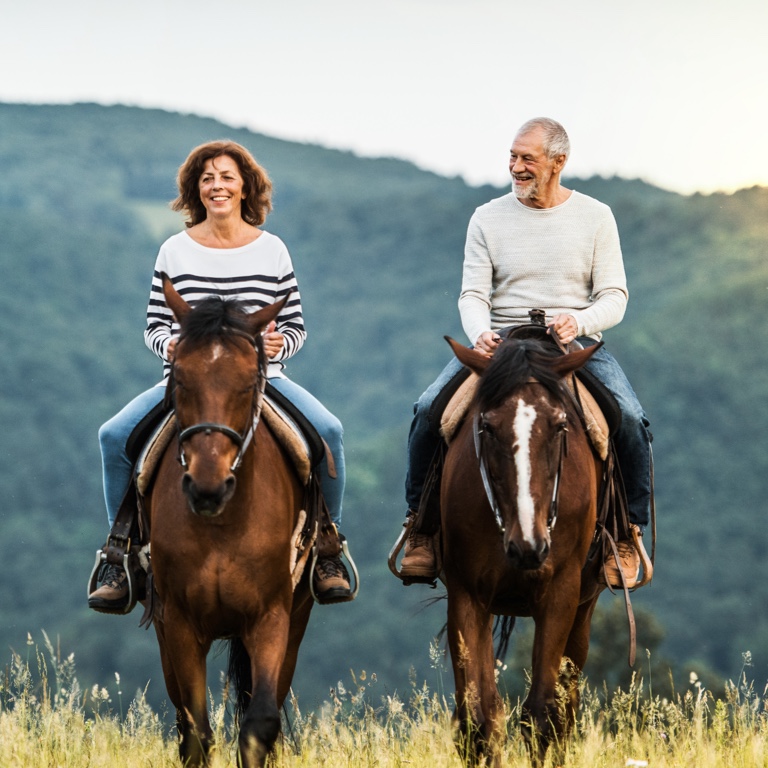 An elderly woman and man, happily riding horses outdoors with mountains in the background.