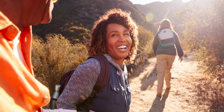 Happy mature woman in the outdoors on a hiking trail, with a fellow female hiker walking ahead in the distance.