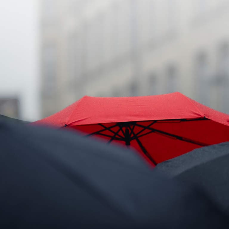 Red umbrella over many black umbrellas against a grey, cloudy sky.