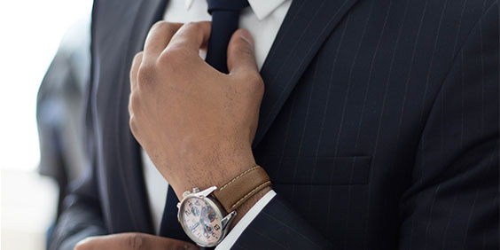 Close-up of a financial adviser getting ready to see his client; fixing his tie, wearing a dark suit, a white shirt and a brown leather strap watch.