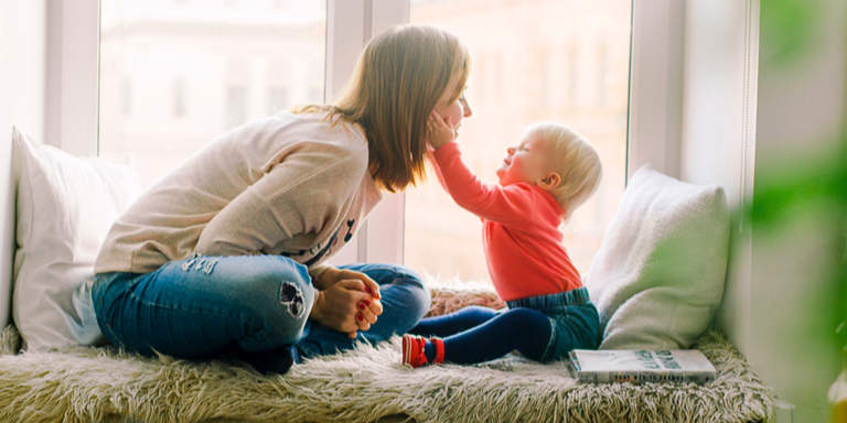 Side view of a happy mother sitting, resting on a bed with her toddler who is laughing and holding his moms face in his hands.