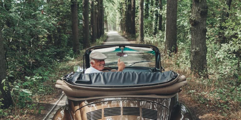An old man drive his restored VW Beetle with the top down through a wooded are, look back at the camera holding his right hand in a thumbs-up sign.