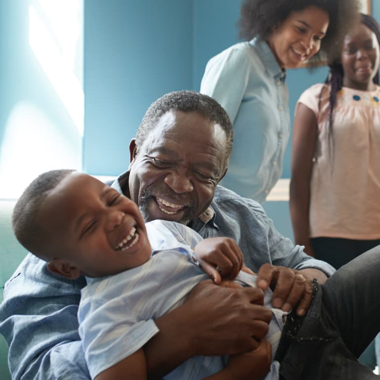 A grandfather tickles and embraces his grandson while his daughter and granddaughter look at them smiling.