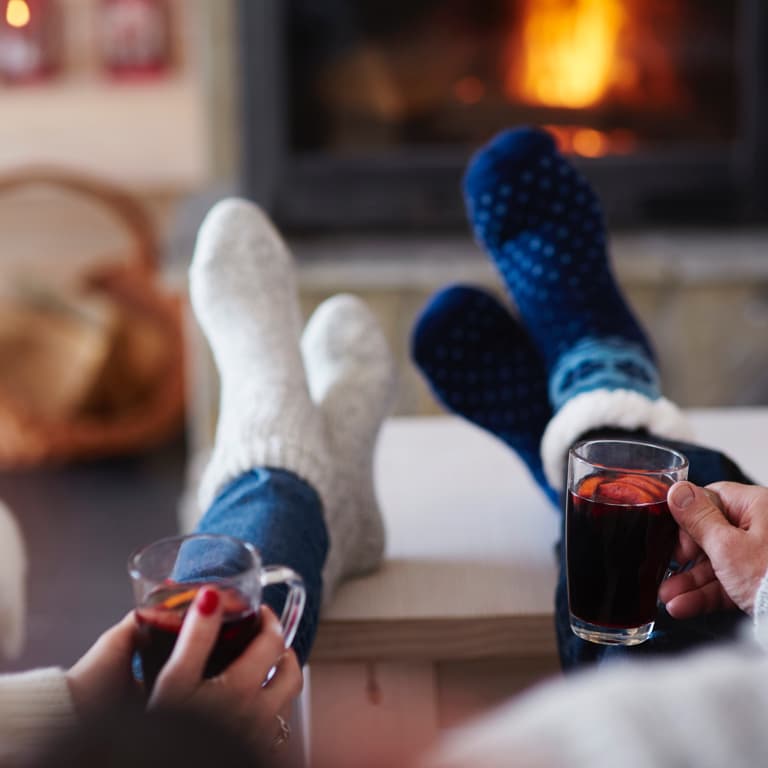 A couple sit in front of a fire with their sock-covered feet on the coffee table, drinking a warm drink.