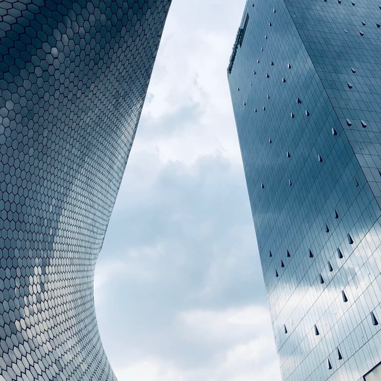 Two modern, abstract high-rise office buildings, with grey blue glass exteriors, amidst a cloudy sky backdrop.