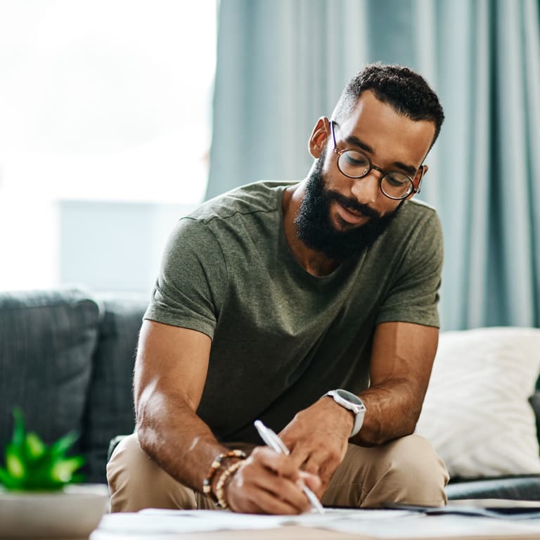 A young muscular man with a beard and glasses, sits on his couch completing his will.